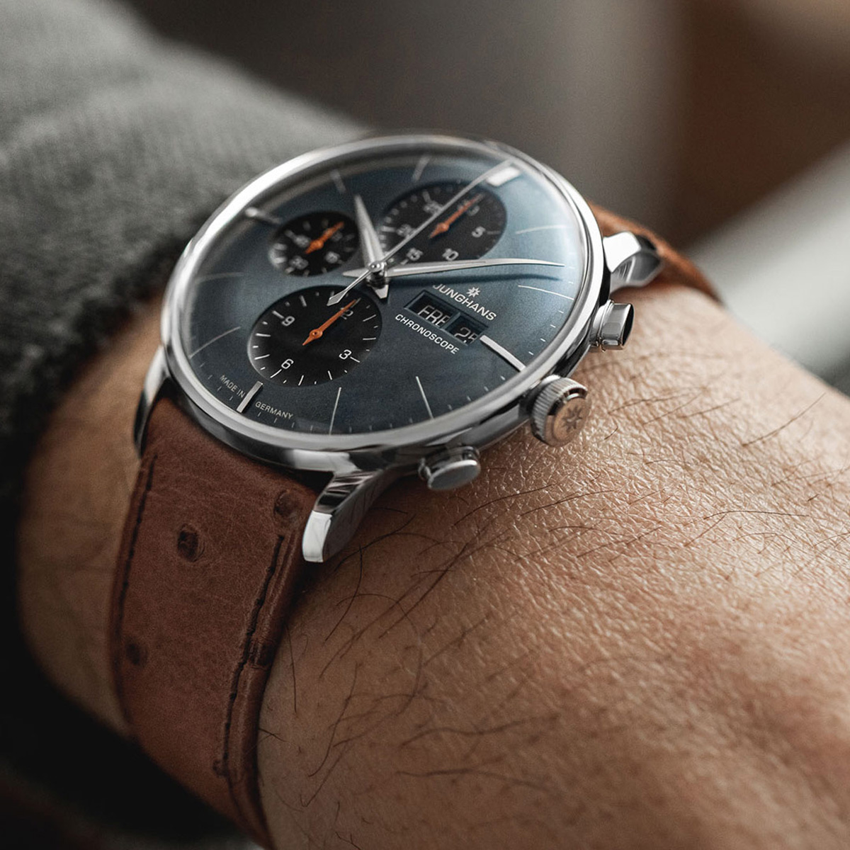 Eine Nahaufnahme des Handgelenks einer Person, die eine Junghans Meister Kollektion Chronographenuhr mit braunem Lederarmband, dunklem Zifferblatt und silbernem Gehäuse trägt. Die Uhr zeigt drei Hilfszifferblätter und orangefarbene Akzente auf den Zeigern.
