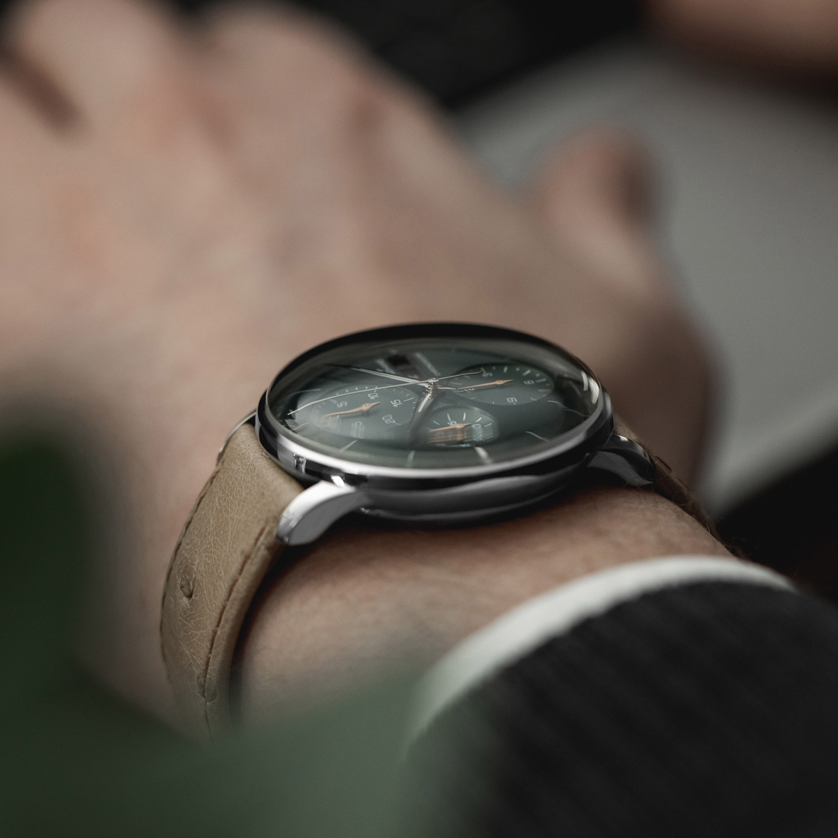JUNGHANS © 2026 Nahaufnahme eines Handgelenks mit einer Junghans Meister Automatik-Armbanduhr mit braunem Lederarmband und dunklem, reflektierendem Zifferblatt. Die Person ist mit einer dunklen Jacke bekleidet und benutzt im Hintergrund eine Tastatur.