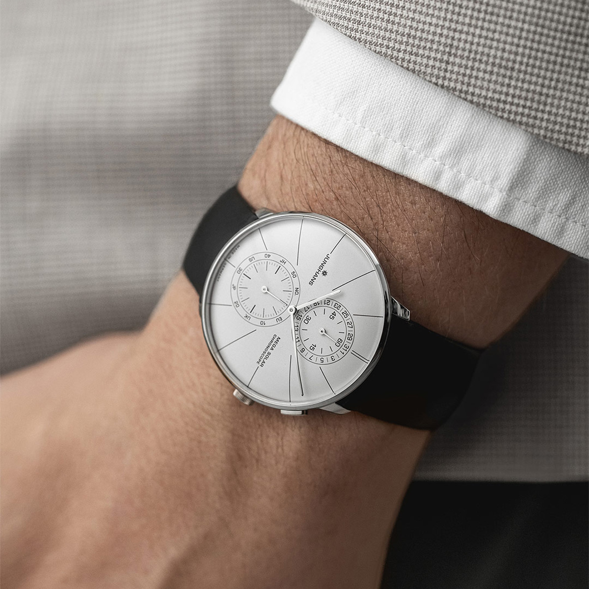 Eine Person, die eine schlichte, moderne Junghans Meister Funk und Solar Armbanduhr mit weißem Zifferblatt, zwei sich überschneidenden Hilfszifferblättern, schwarzem Armband und minimalistischem Design trägt, bekleidet mit einer hellgrau gemusterten Jacke und einem weißen Hemd.