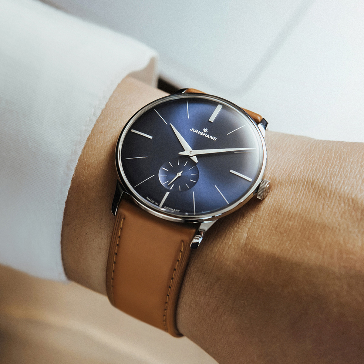 JUNGHANS © 2026 Eine Person mit einer Junghans Meister Handaufzug Armbanduhr mit braunem Lederarmband und blauem Zifferblatt, bekleidet mit einem weißen Hemd mit Manschetten.