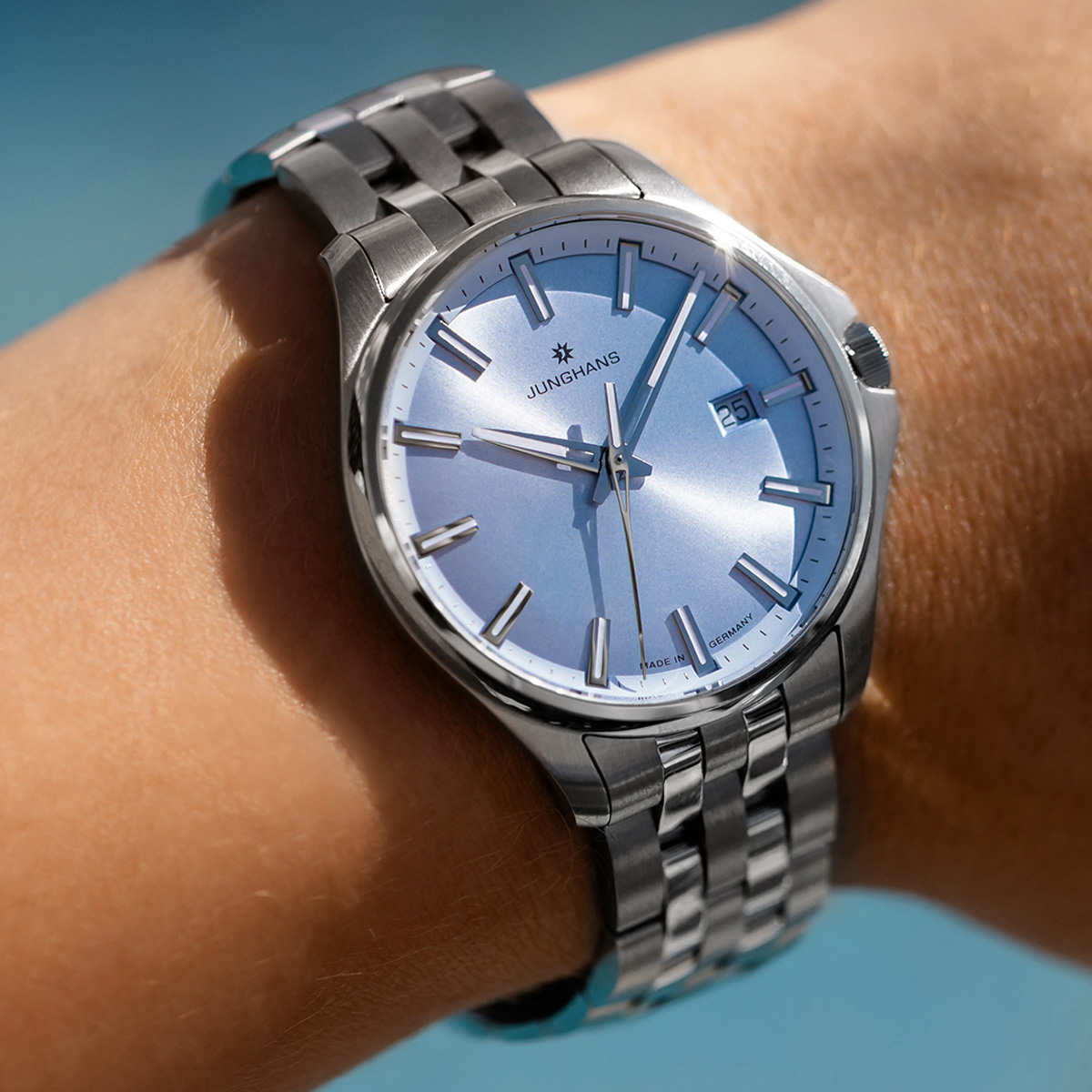 JUNGHANS © 2026 Nahaufnahme einer silbernen Junghans Meister Quarz-Armbanduhr mit metallisch blauem Zifferblatt und Metallband am Handgelenk einer Person, die die Uhrzeit 10:09 und das Datum 25 anzeigt, vor einem verschwommenen blauen Hintergrund.