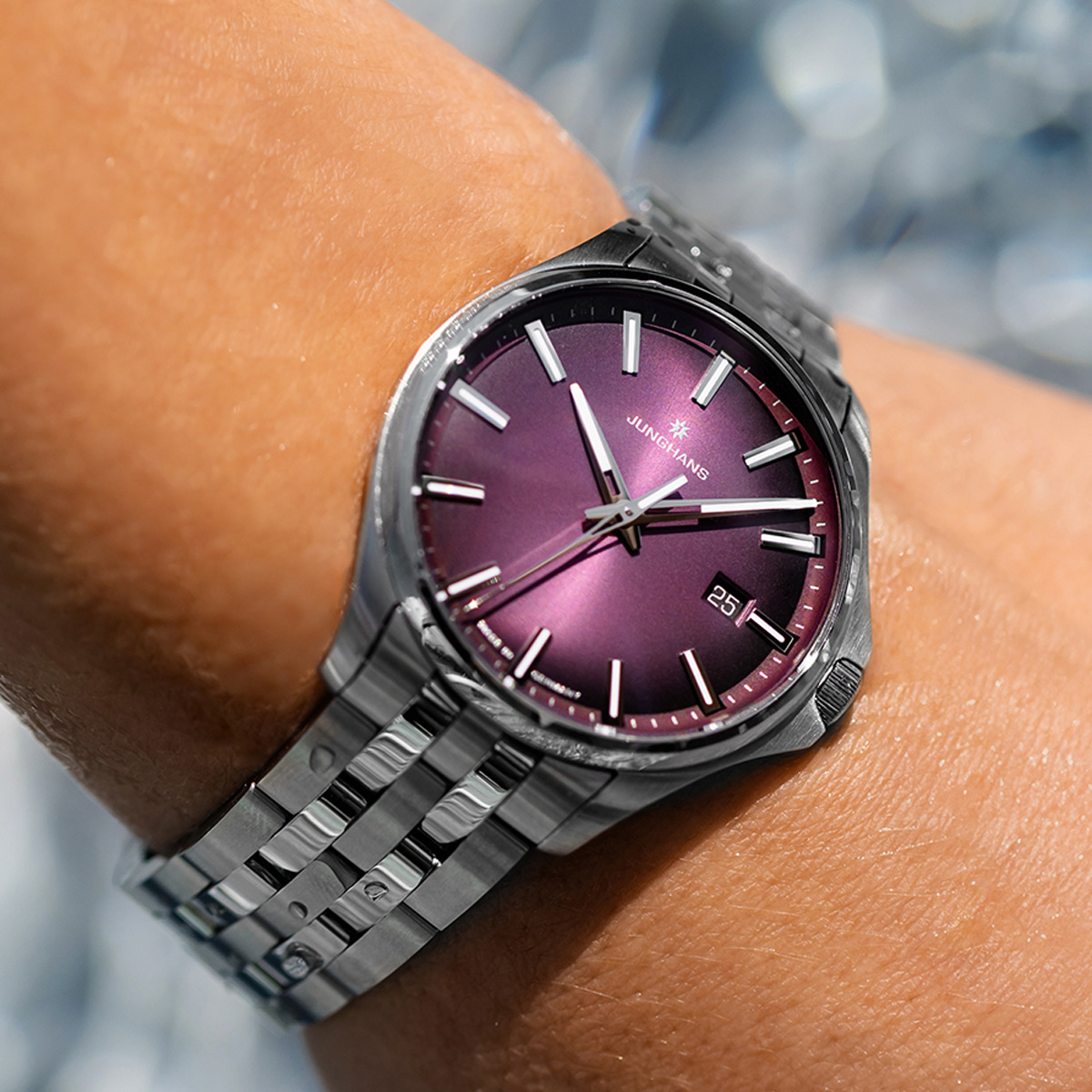 JUNGHANS © 2026 Nahaufnahme einer Junghans Meister Quarz-Armbanduhr mit lilafarbenem Metallzifferblatt und Datumsfenster, die am Handgelenk einer Person getragen wird. Das silberne Metallband funkelt im Sonnenlicht und unterstreicht das elegante Design der Uhr.