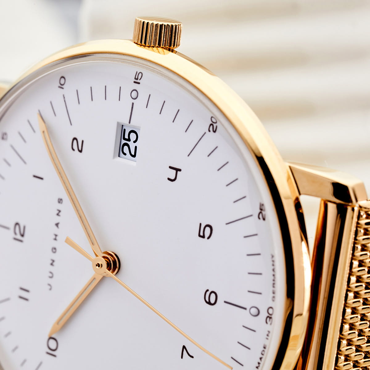 JUNGHANS © 2026 Nahaufnahme einer goldenen Junghans Max Bill Armbanduhr mit weißem Zifferblatt, minimalistischen schwarzen Indexen, einem Datumsfenster mit der Zahl "25" und einem goldenen Netzarmband. Die Uhr ist leicht angewinkelt, was ihr elegantes Design unterstreicht.