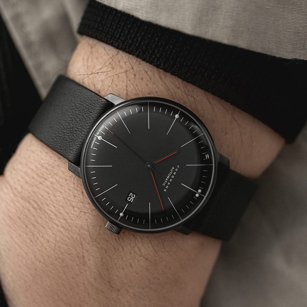 Nahaufnahme des Handgelenks einer Person, die eine minimalistische schwarze Junghans Max Bill Armbanduhr mit schwarzem Lederarmband, schwarzem Zifferblatt, weißen Markierungen und einem roten Sekundenzeiger trägt. Das Datumsfenster zeigt "25" an. Die Person trägt eine beigefarbene Jacke.