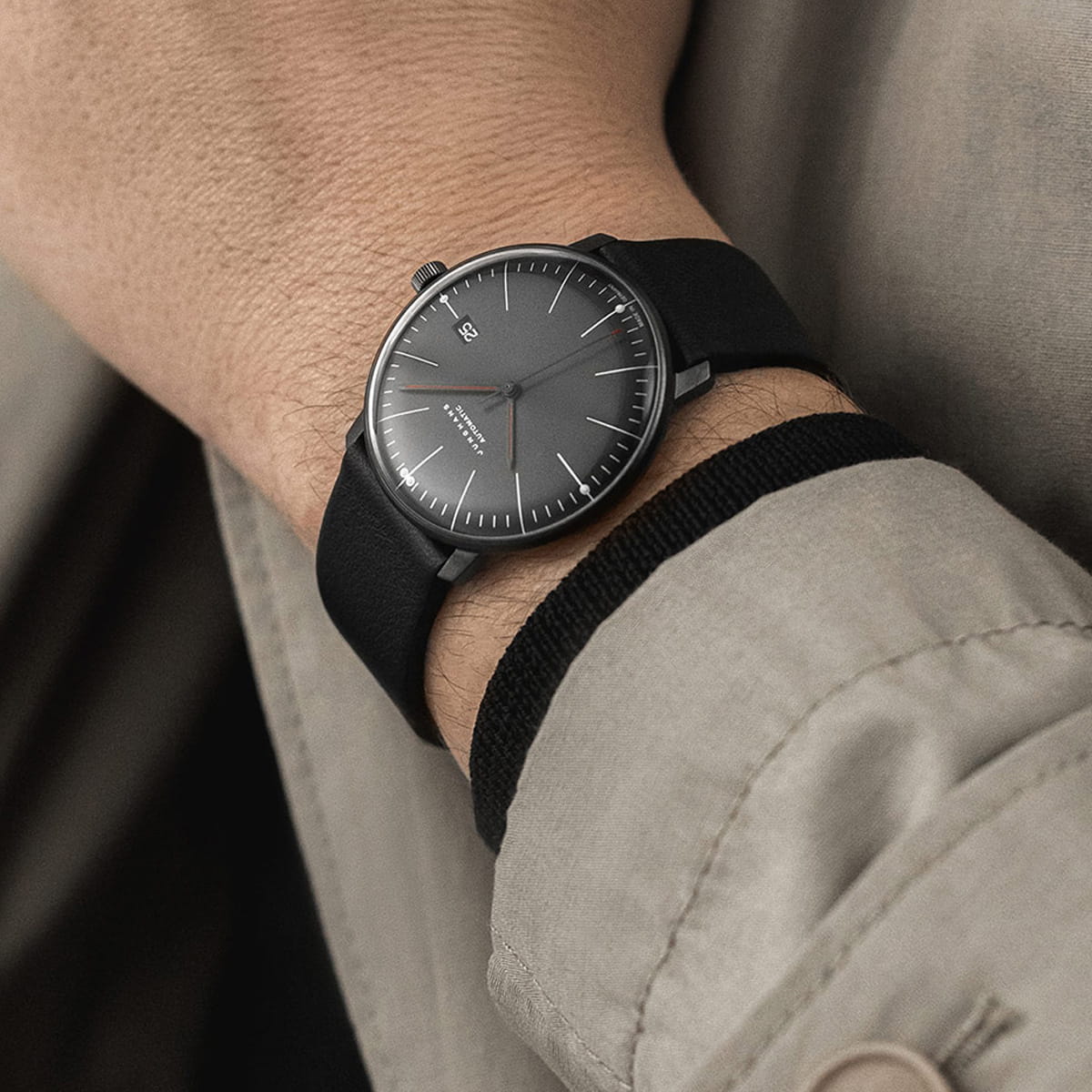 Eine Person, die eine minimalistische schwarze Junghans Max Bill Automatik-Armbanduhr mit dunkelgrauem Zifferblatt und weißen Markierungen trägt, bekleidet mit einem hellbeigen Mantel.