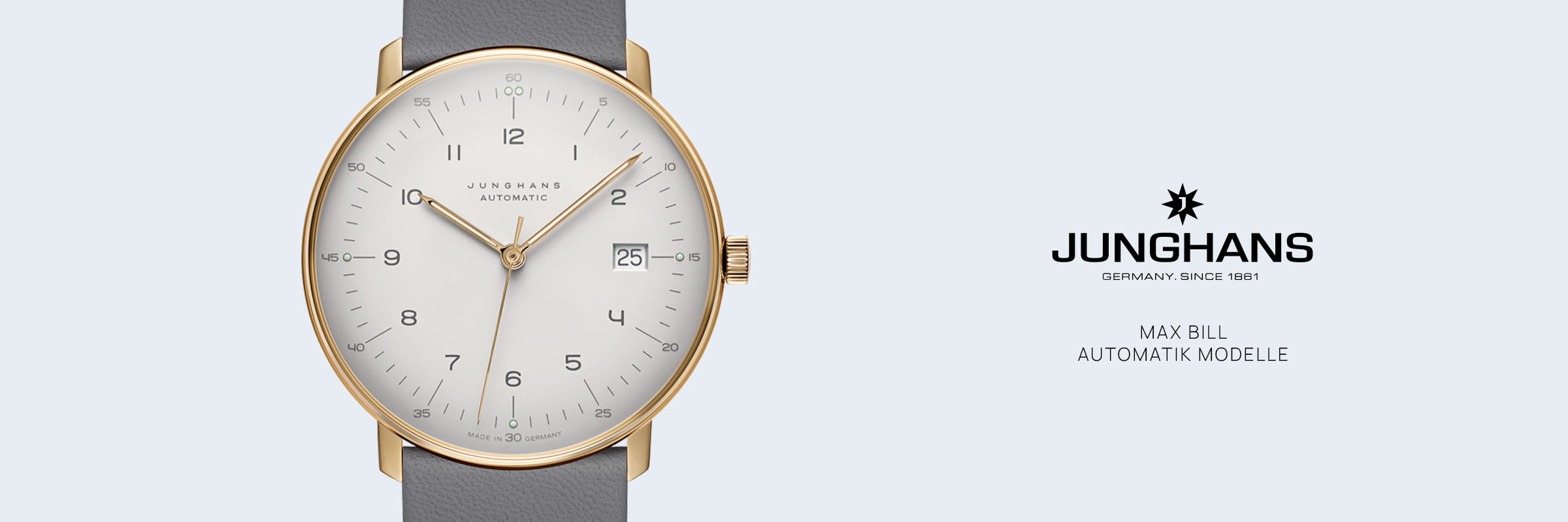 Eine Junghans Max Bill Automatik Armbanduhr mit goldenem Gehäuse, weißem Zifferblatt, schwarzen Indexen und Datumsanzeige bei 3 Uhr an einem grauen Lederarmband, das neben dem Junghans-Logo und der Modellbezeichnung auf hellem Grund steht.