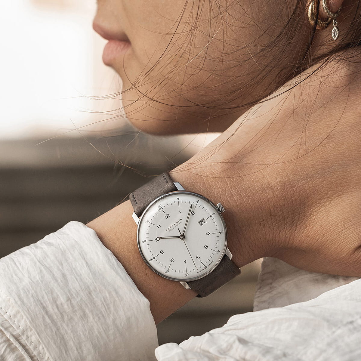 JUNGHANS © 2026 Eine Nahaufnahme des Handgelenks einer Frau, die eine minimalistische Junghans Max Bill Silberuhr mit weißem Zifferblatt und grauem Lederarmband trägt. Sie ist mit einem weißen Hemd bekleidet und trägt goldene Ohrringe im Ohr.