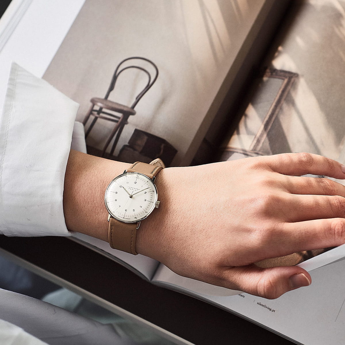 Eine Person, die einen Junghans Max Bill Handaufzug mit hellbraunem Lederarmband und weißem Zifferblatt trägt, stützt ihre Hand auf ein aufgeschlagenes Buch. Im Hintergrund vervollständigen ein verschwommener Stuhl und weiche Schatten die heitere Szene.