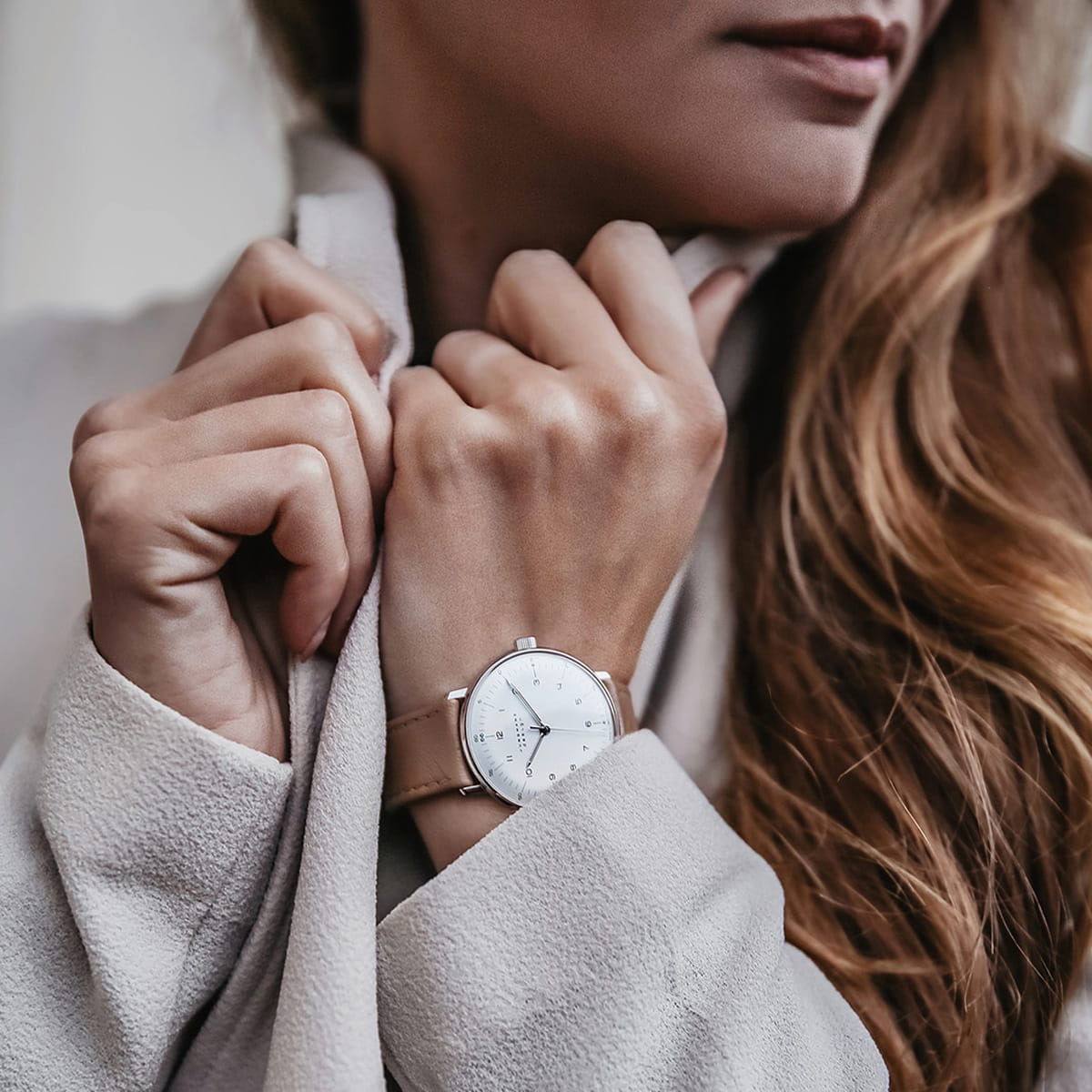 Eine Frau mit langem, hellbraunem Haar rückt den Kragen eines hellbeigen Mantels zurecht und präsentiert eine minimalistische silberne Armbanduhr von Junghans Max Bill Handaufzug mit weißem Zifferblatt und hellbraunem Lederarmband an ihrem linken Handgelenk.