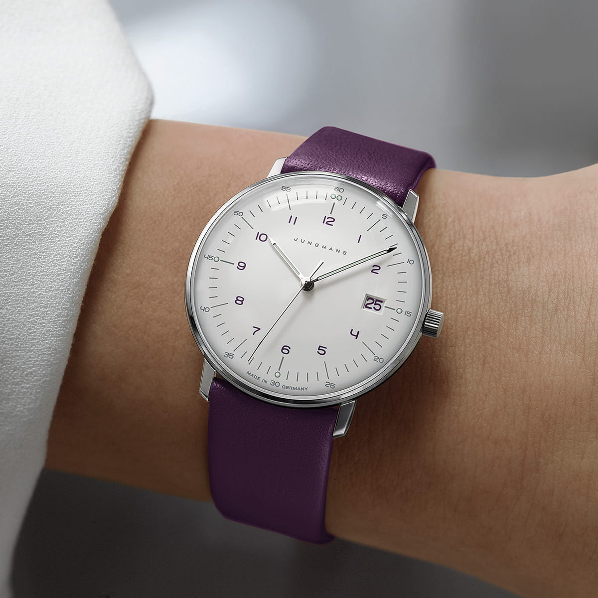 JUNGHANS © 2026 Eine minimalistische Junghans Max Bill Quarz Armbanduhr mit weißem Zifferblatt, silbernem Gehäuse, lilafarbenem Lederband und schwarzen Ziffern wird am Handgelenk einer Person getragen. Die Uhr zeigt das Datum an und besticht durch ein modernes, klares Design.