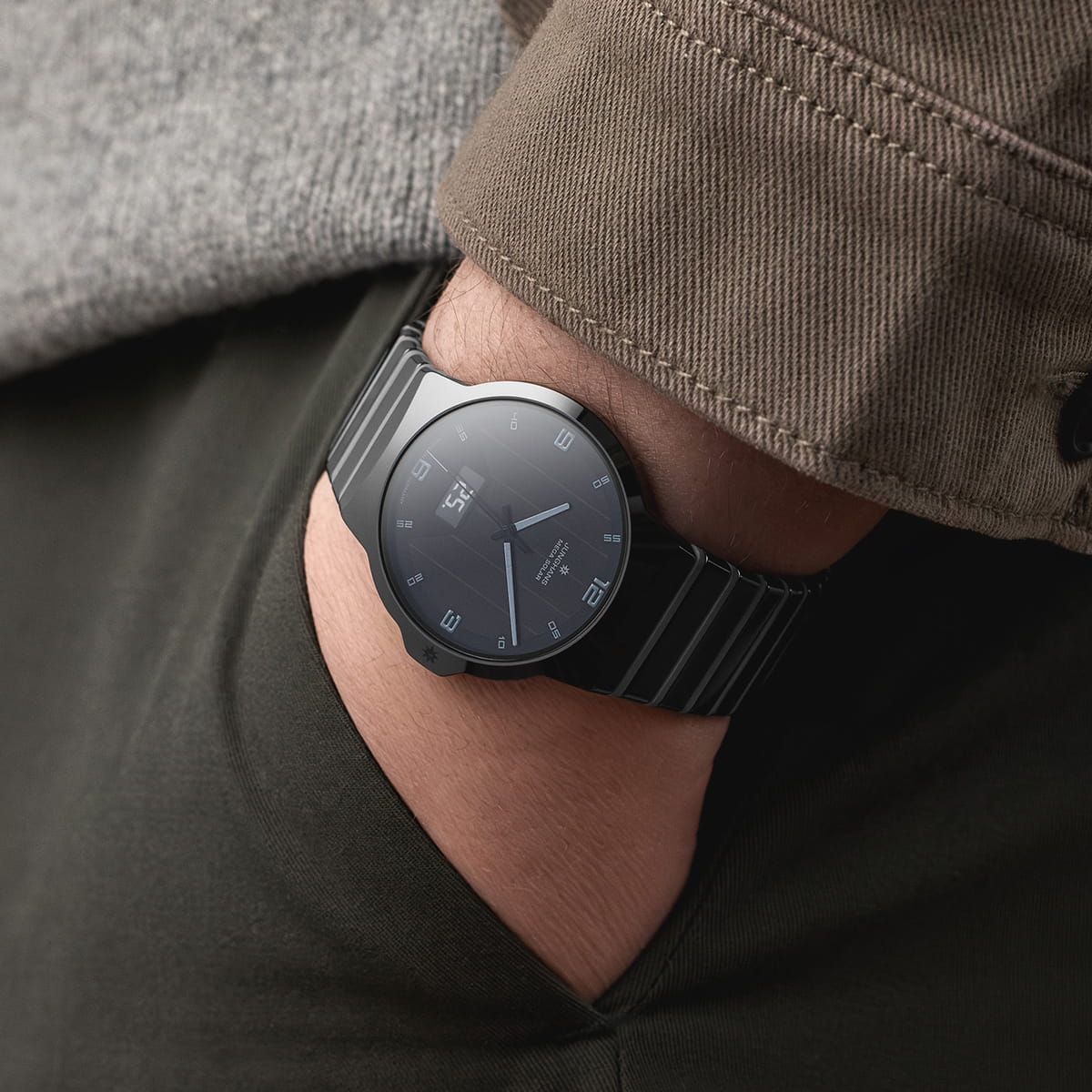 Juwelier-Wagner-Madler-Schmuck-und-Uhren-Mainz-Junghans-Kollektion-Performance-Ansicht-1-Force Eine Person, die eine dunkle, schlichte Junghans Performance-Armbanduhr mit rundem Zifferblatt und dünnen Stundenmarkierungen trägt, hat die Hand in der Tasche einer dunklen Hose, die mit einer braunen Jacke und einem grauen Pullover bekleidet ist.