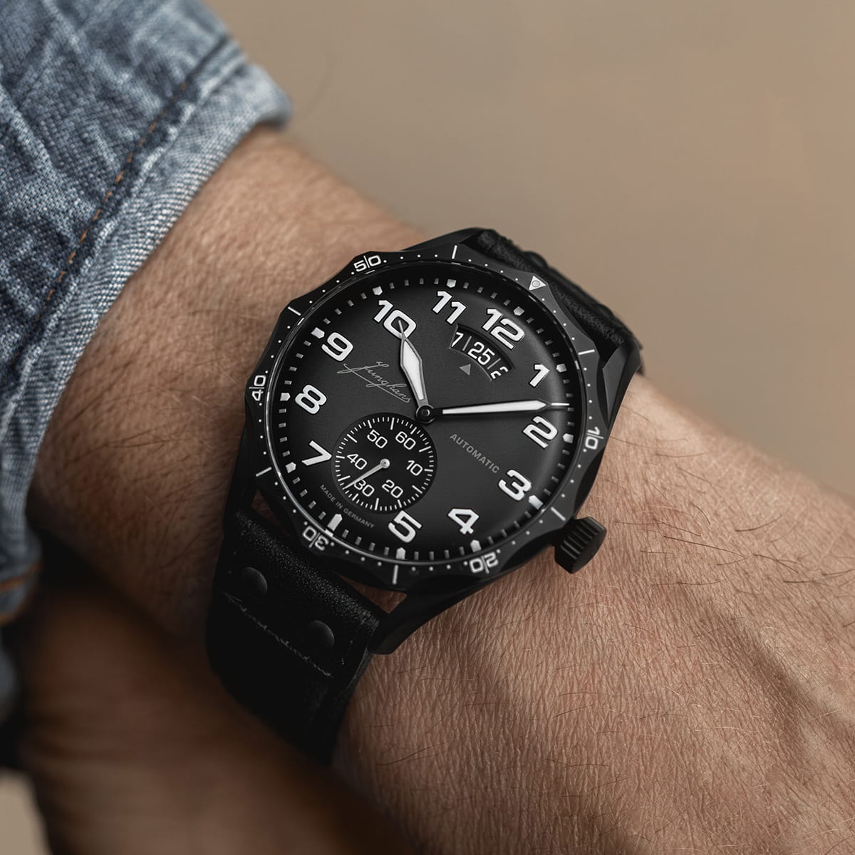 Nahaufnahme des Handgelenks einer Person, die eine schwarze analoge Junghans Pilot-Armbanduhr mit weißen Ziffern, einer Datumsanzeige und einem schwarzen Lederarmband trägt. Die Person ist mit einem Jeansärmel bekleidet.