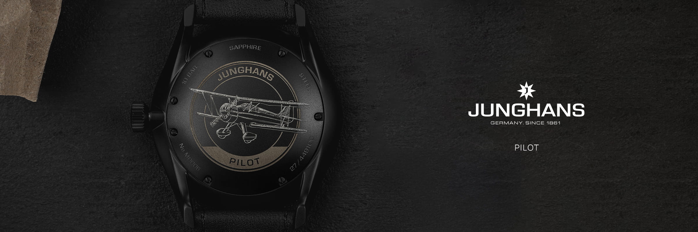 Nahaufnahme der Rückseite einer Junghans-Fliegeruhr mit eingraviertem Doppeldecker. Auf der rechten Seite heben sich das Junghans-Pilot-Logo und der Text "GERMANY. SINCE 1861" und "PILOT" heben sich von einem dunklen Hintergrund ab.