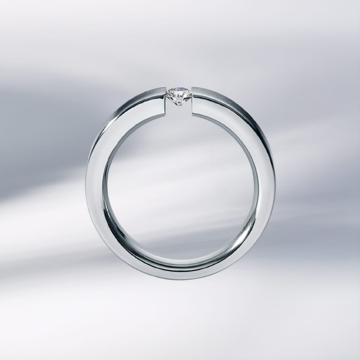 Ein minimalistischer silberner Humphrey One Ring mit offenem, kreisförmigem Design und einem einzelnen Diamanten in der Lücke wird vor einem weichen, verschwommenen, hellgrauen Hintergrund gezeigt.