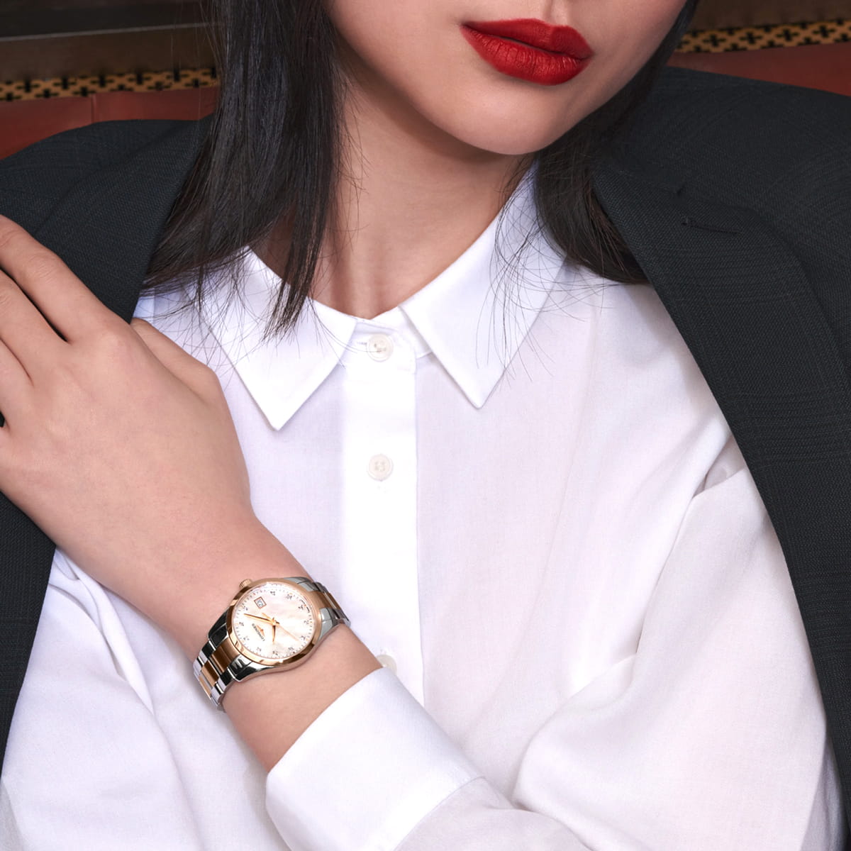 Eine Frau, die ein weißes Hemd mit Kragen, einen dunklen Blazer und roten Lippenstift trägt, stützt ihre Hand auf ihre Schulter und präsentiert elegant eine silberne Longines Conquest Classic-Armbanduhr mit weißem Zifferblatt an ihrem linken Handgelenk.