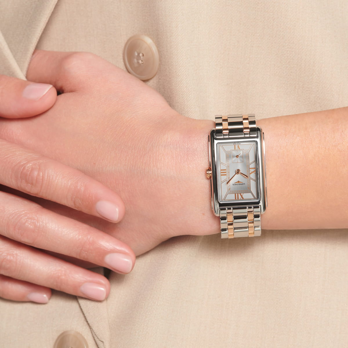LONGINES © 2026 Eine Person, die eine rechteckige Longines DolceVita-Armbanduhr in Gold und Silber mit weißem Zifferblatt und römischen Ziffern trägt, bekleidet mit einer beigefarbenen, geknöpften Jacke und ordentlich manikürten Nägeln.