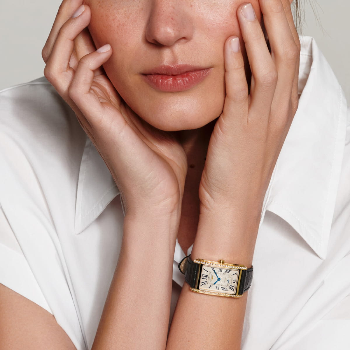 LONGINES © 2026 Eine Frau in einem weißen Hemd mit Kragen stützt ihr Gesicht in die Hände und trägt eine rechteckige Longines DolceVita-Armbanduhr aus Gold mit schwarzem Lederband an ihrem linken Handgelenk. Auf dem Zifferblatt sind elegante römische Ziffern zu sehen.