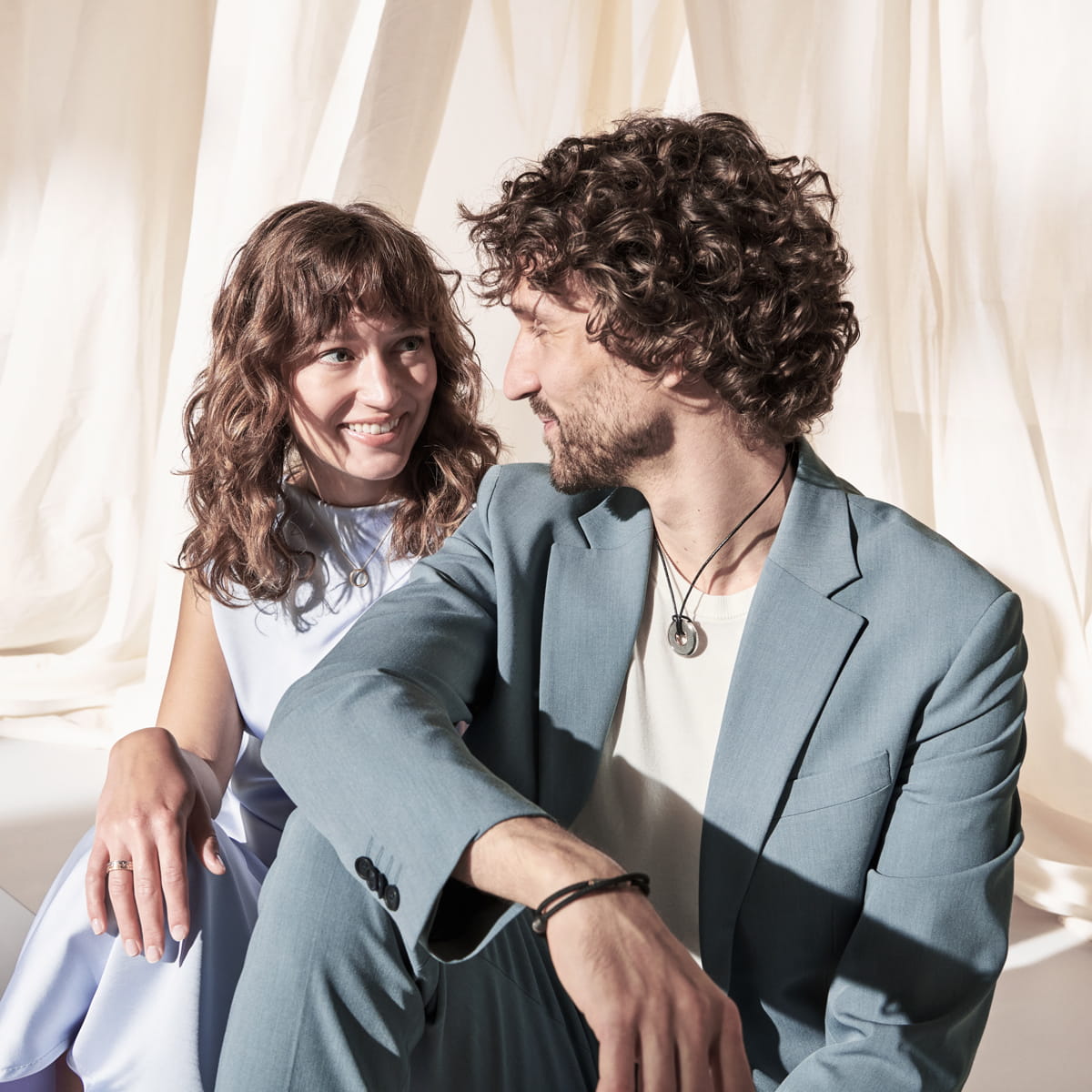 Eine Frau und ein Mann sitzen dicht beieinander und lächeln sich herzlich an. Der Mann trägt einen hellblauen Anzug von Meister, die Frau ein helles Kleid, und sie sind von weichen, cremefarbenen Vorhängen umgeben.