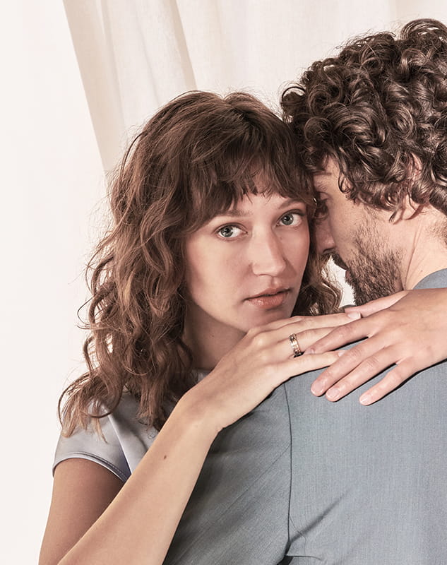 Eine Frau mit gewelltem braunem Haar umarmt einen Mann mit lockigem Haar, der ruhig in die Kamera schaut. Sie legt ihre Hand, die mit einem Meisterring geschmückt ist, auf seine Schulter, während er sich vom Objektiv abwendet.