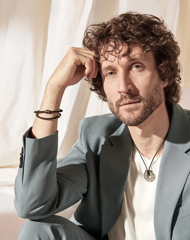 Ein Mann mit lockigem braunem Haar und Bart sitzt nachdenklich da, trägt einen hellblauen Meister-Anzug, ein weißes T-Shirt, eine schwarze Halskette und ein Armband und stützt sich mit dem Ellbogen auf sein Knie vor einem hellen, sanft beleuchteten Hintergrund.