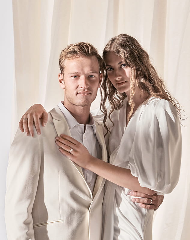 Ein Paar in weißer Meisterkleidung steht dicht beieinander vor weißen Vorhängen, wobei die Frau den Arm um die Schulter des Mannes legt und beide in die Kamera schauen.
