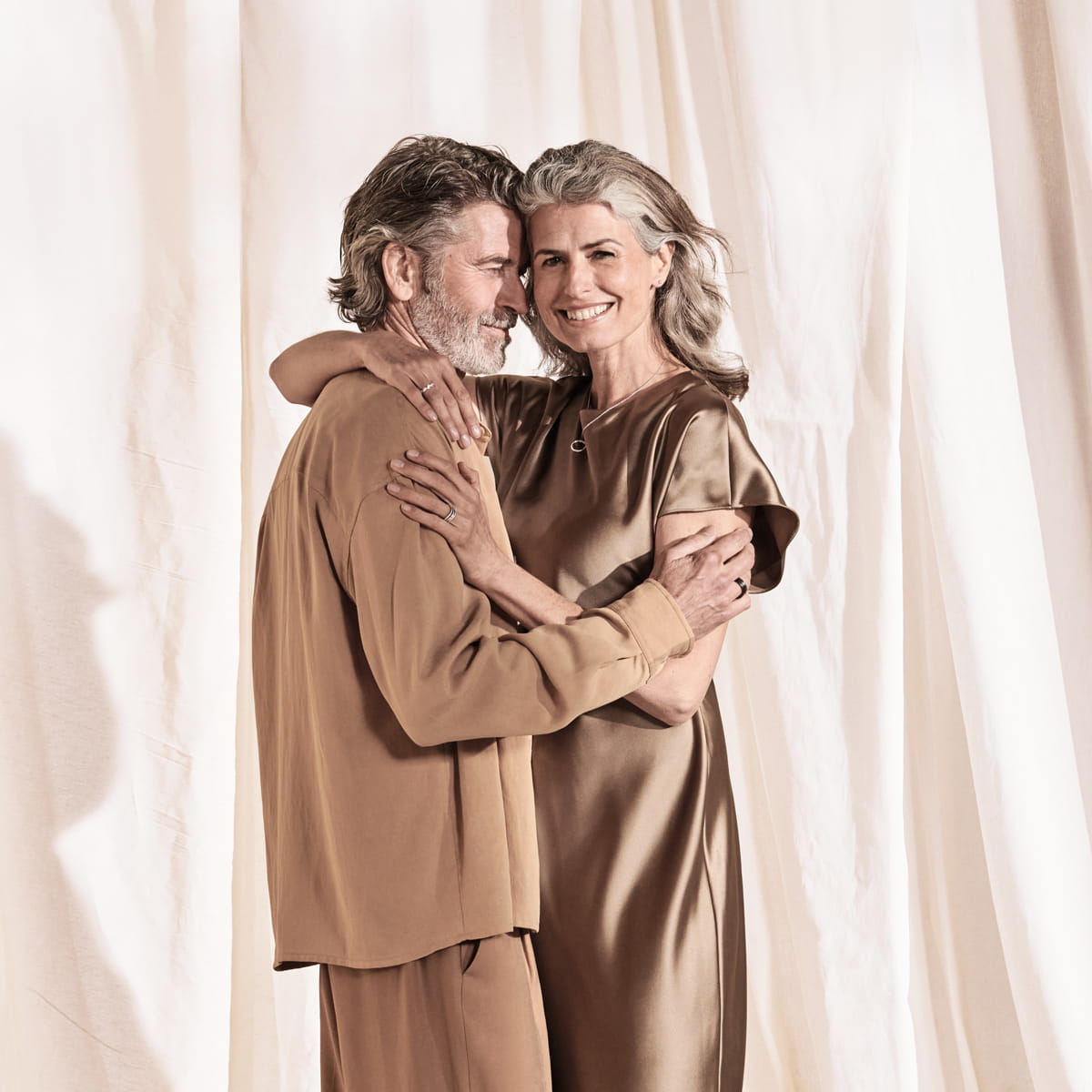 Ein lächelndes älteres Paar mit grauem Haar, das passende hellbraune Outfits trägt, umarmt sich vor leichten, fließenden Stoffkulissen und strahlt ein Meistergefühl von zeitloser Eleganz und Verbundenheit aus.