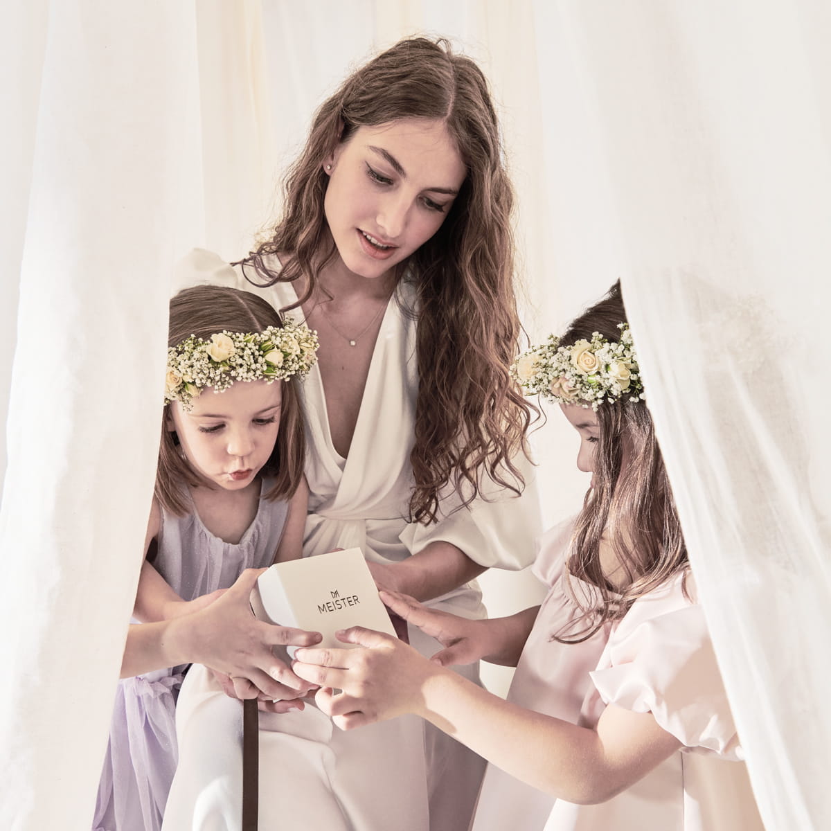 Eine Frau in einem weißen Kleid sitzt zwischen zwei jungen Mädchen mit Blumenkränzen und pastellfarbenen Kleidern, die gemeinsam eine weiße MEISTER-Geschenkbox bewundern, umgeben von weichen, weißen Meister-Vorhängen.
