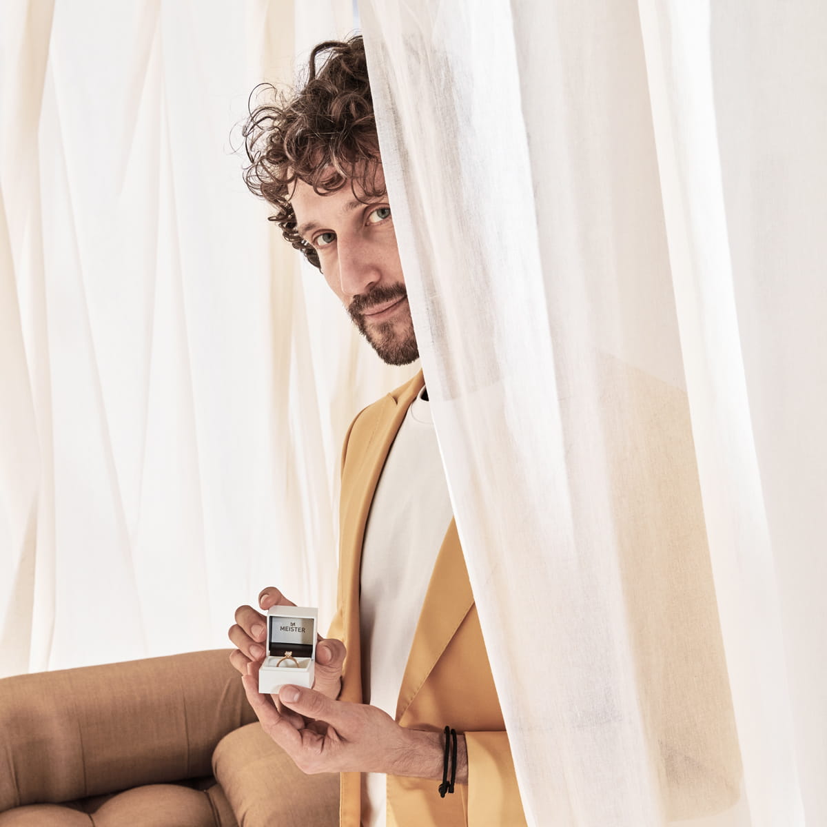 Ein Mann mit lockigem Haar und Bart steht hinter weißen Vorhängen, trägt einen gelben Meister-Blazer und hält ein kleines quadratisches Parfümfläschchen. Neben ihm ist ein brauner, gepolsterter Stuhl teilweise sichtbar.