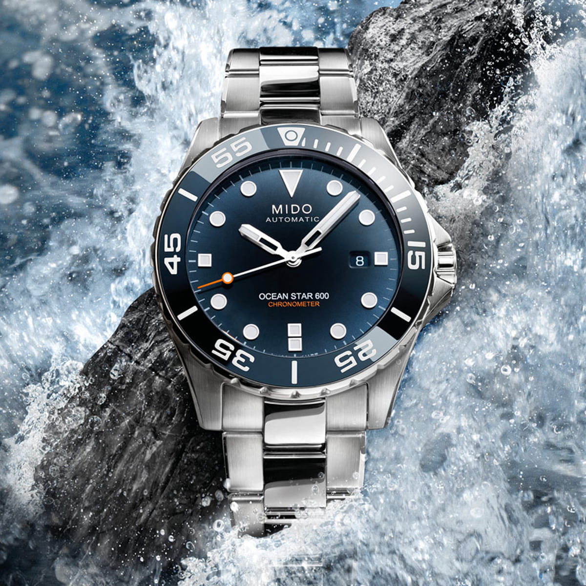 MIDO © 2026 Eine silberne Mido Ocean Star 600 Automatik-Armbanduhr mit blauem Zifferblatt und Edelstahlarmband wird vor dem Hintergrund der plätschernden Wellen des Ozeans präsentiert und unterstreicht die berühmte Widerstandsfähigkeit und Wasserdichtigkeit der Marke Mido.