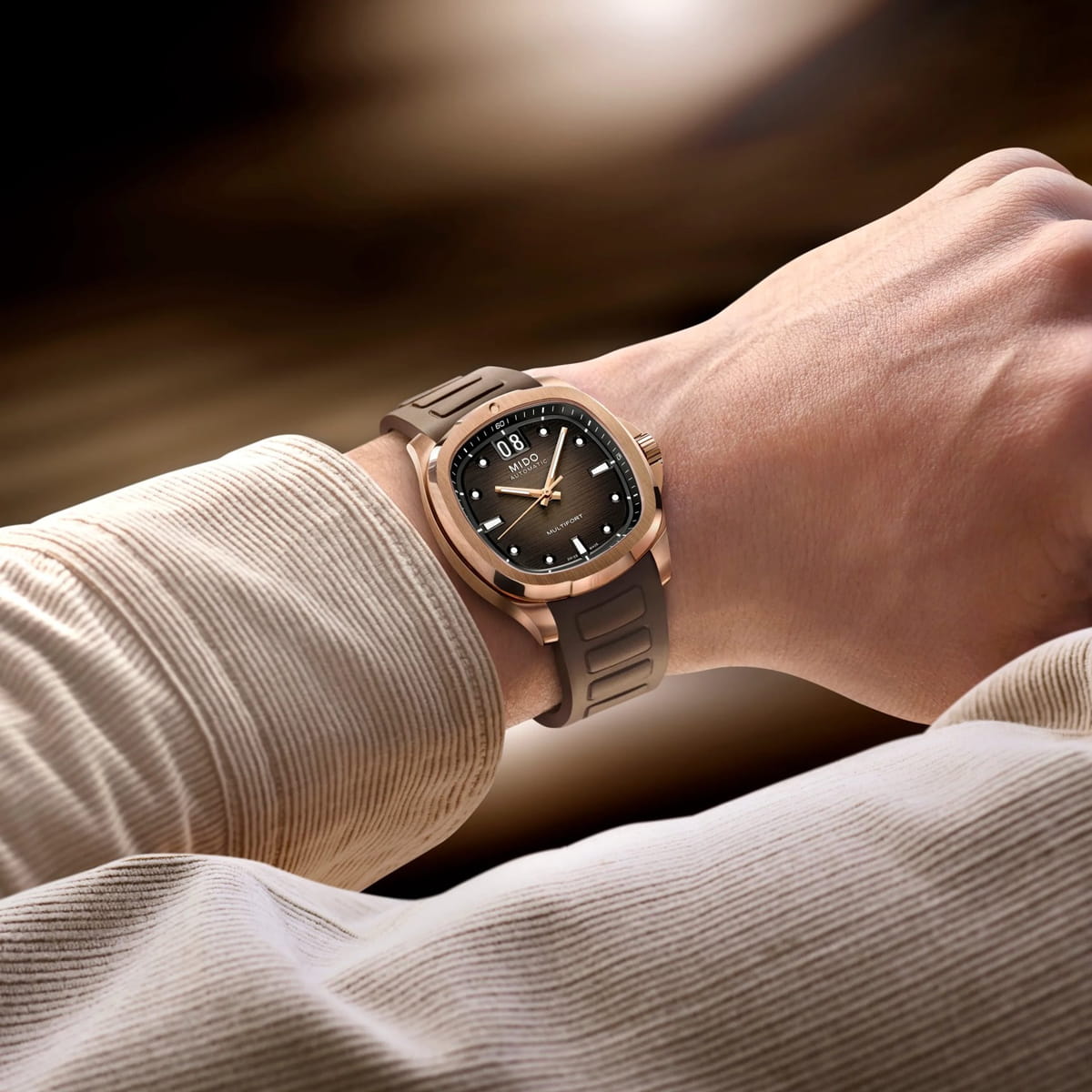 MIDO © 2026 Eine Person trägt eine stilvolle braun-goldene Mido-Armbanduhr mit rundem Zifferblatt, dunklem Ziffernblatt und weißen Stundenmarkierungen. Die Person ist mit einem hellbeigen, gerippten langärmeligen Hemd bekleidet. Der Hintergrund ist sanft verschwommen.