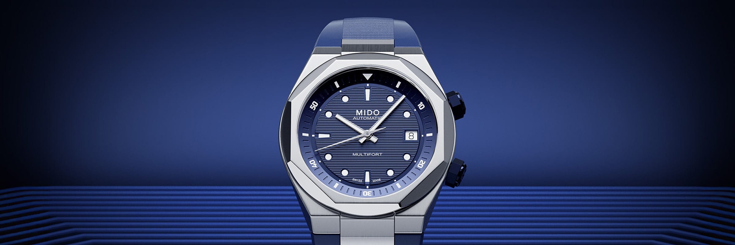 MIDO © 2026 Eine blau-silberne Mido-Armbanduhr mit strukturiertem blauem Zifferblatt, weißen Stundenmarkierungen und einem Datumsfenster auf blauem Hintergrund mit horizontalen Linien.