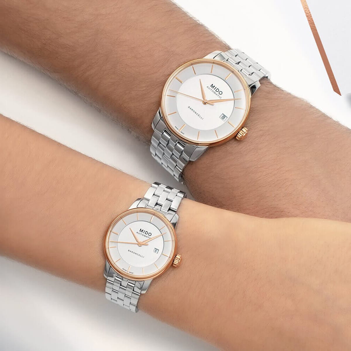 Zwei Handgelenke mit passenden silbernen Mido Baroncelli-Armbanduhren mit weißen Zifferblättern, roségoldenen Lünetten und Metallgliederbändern. Die Uhren tragen den Markennamen "Mido" und haben ein Datumsfenster auf der 3-Uhr-Position.