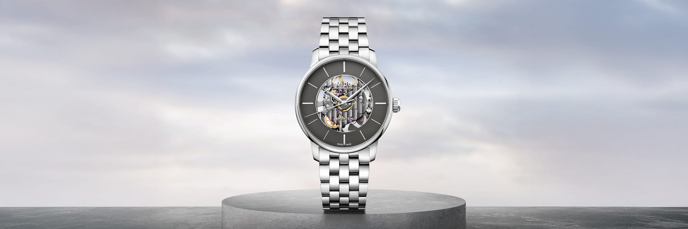 Eine silberne Luxus-Armbanduhr von Mido Baroncelli mit Metallgliederband und offenem mechanischem Uhrwerk wird auf einem runden Sockel vor einem weichen, wolkenverhangenen Hintergrund präsentiert.