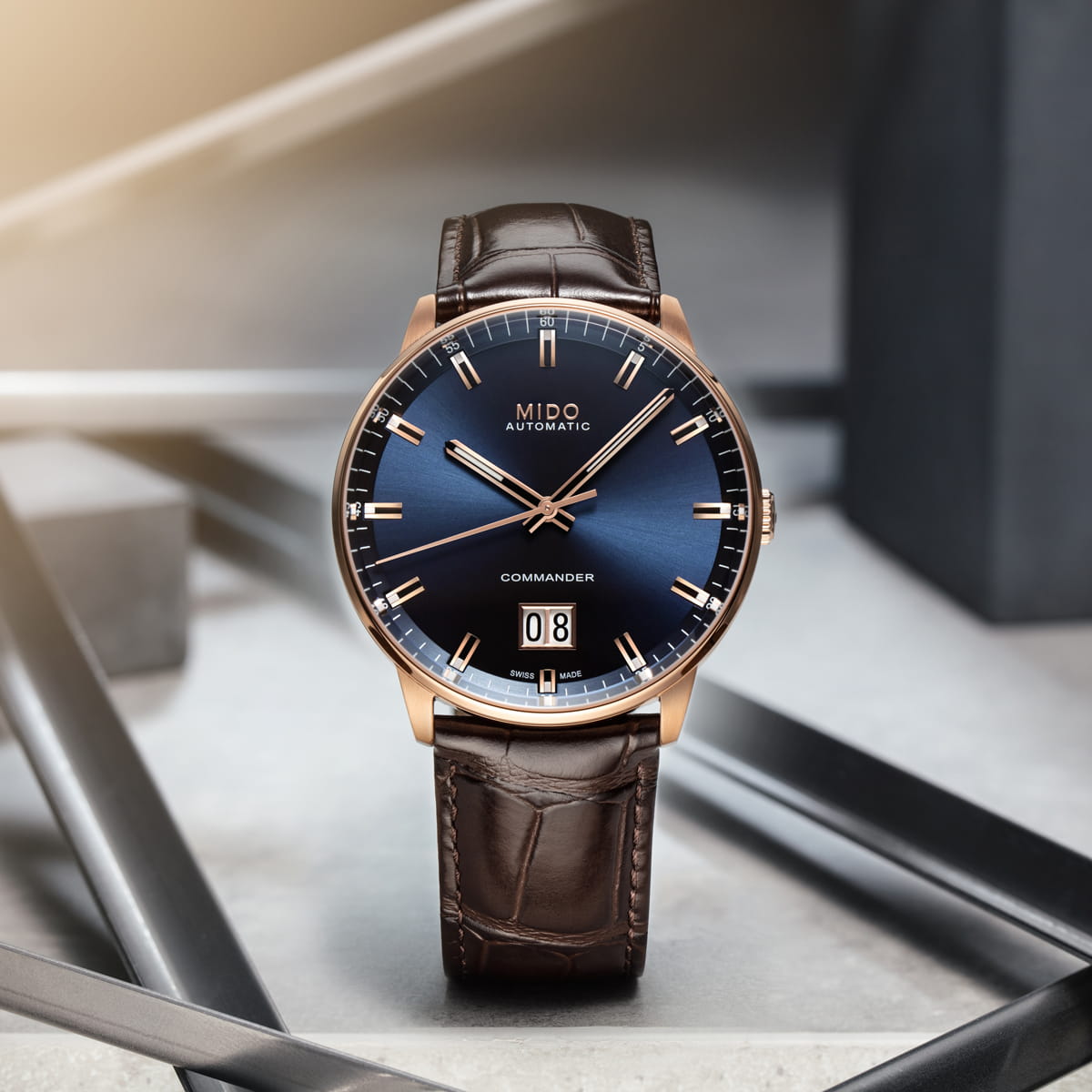 Die Mido Commander mit blauem Verlaufszifferblatt, goldenem Gehäuse und braunem Lederarmband verfügt über eine große Datumsanzeige bei 6 Uhr, die auf einer modernen, geometrischen Oberfläche mit weicher Beleuchtung für einen raffinierten Look sorgt.