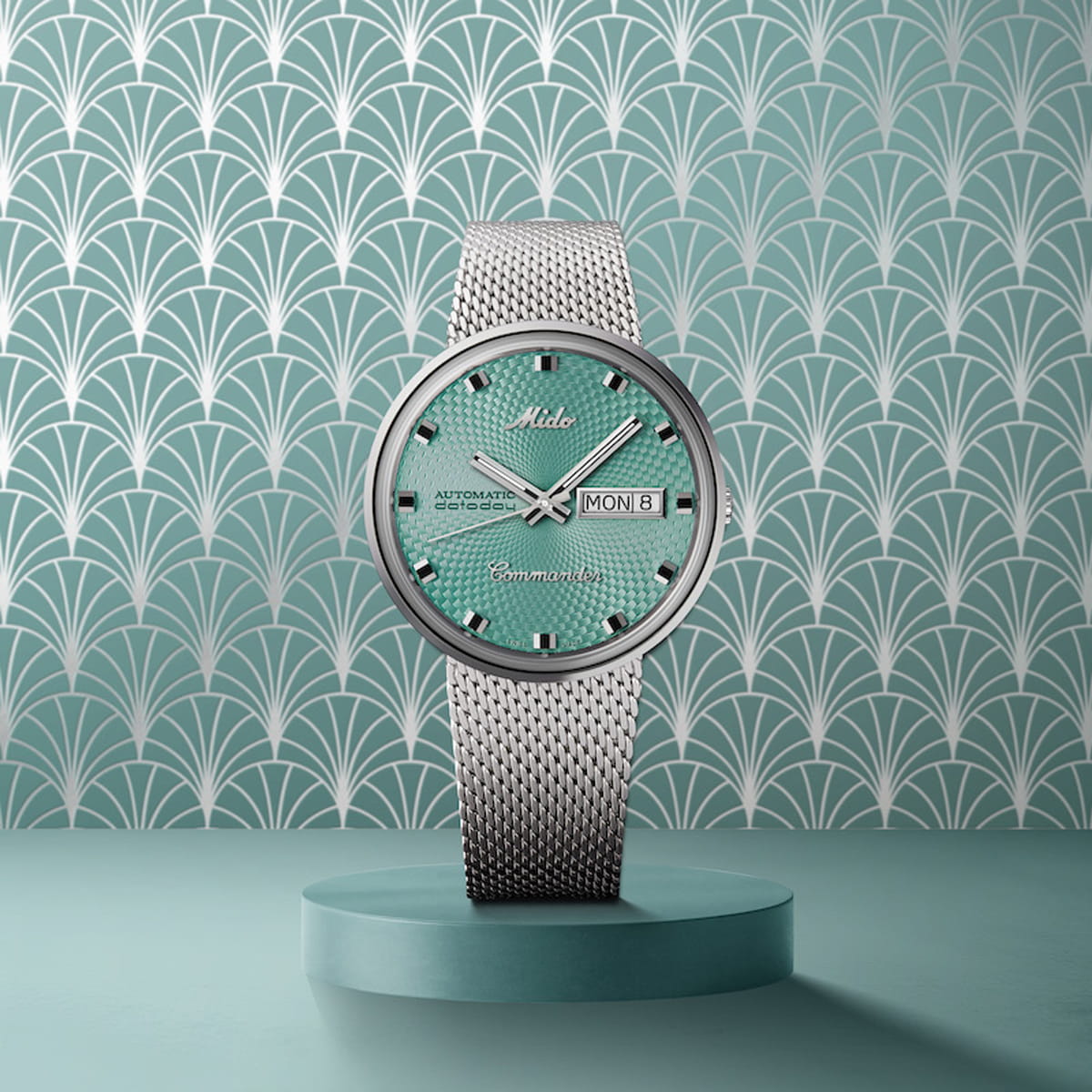 Eine silberne Mido Commander-Armbanduhr mit rosafarbenem, strukturiertem Zifferblatt, silbernen Stundenmarkierungen und einem Netzband steht aufrecht auf einem rosafarbenen Sockel vor einem geometrischen, rosafarbenen und weiß gemusterten Hintergrund. Das Zifferblatt zeigt den Tag und das Datum an.