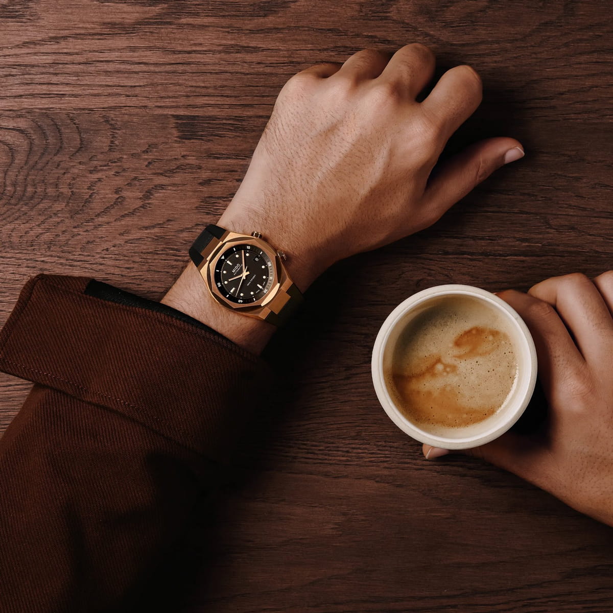 Eine Person, die eine braune Jacke und eine Mido Multifort-Armbanduhr trägt, stützt sich mit einer Hand auf einen Holztisch und hält mit der anderen Hand eine Tasse Kaffee.