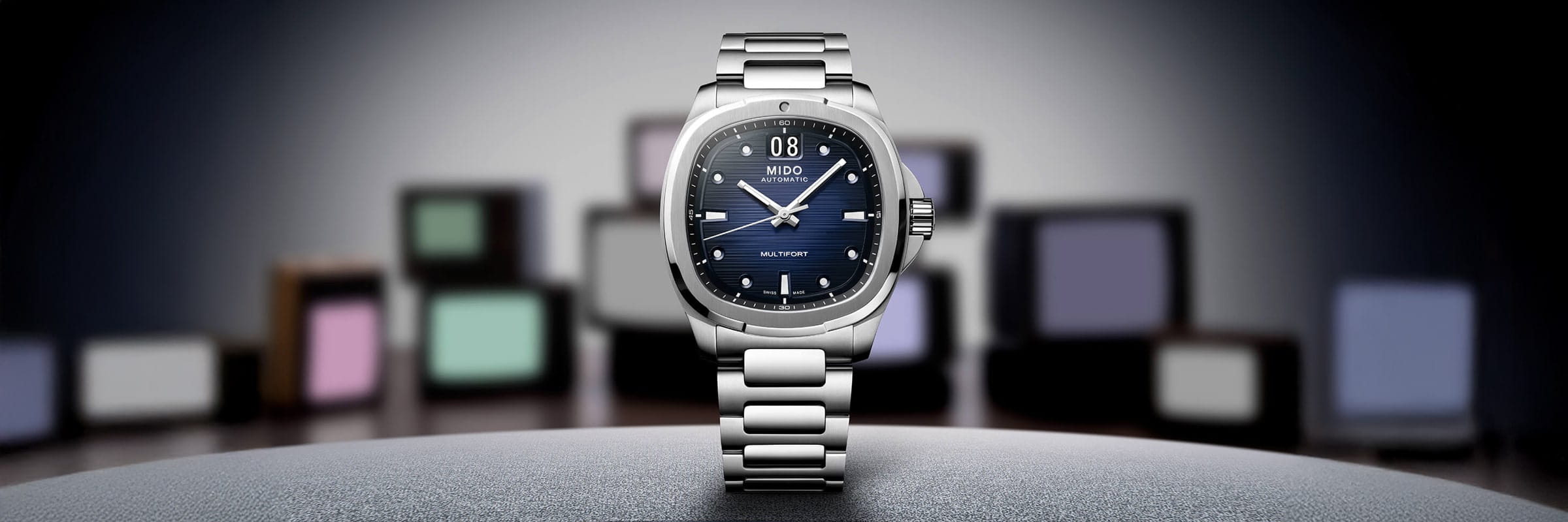 Eine silberne Mido-Multifort-Armbanduhr mit blauem Ziffernblatt steht aufrecht auf einer glatten Oberfläche, mit unscharfen, bunten, quadratischen Objekten im Hintergrund und fokussierter Beleuchtung, die die Uhr hervorhebt.