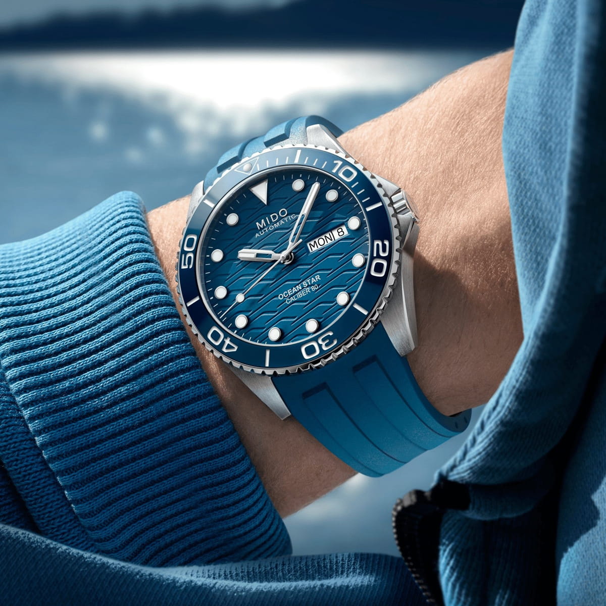 Nahaufnahme eines Handgelenks mit einer auffälligen Mido Ocean Star Taucheruhr mit blauem Kautschukarmband, die über einem blauen Ärmel getragen wird, vor einem unscharfen Wasserhintergrund.