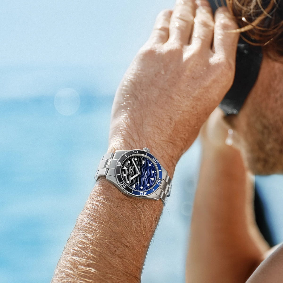 Eine Person, die eine Mido Ocean Star-Armbanduhr mit blauer und schwarzer Lünette trägt, steht an einem sonnigen Tag in der Nähe des Wassers und hält ein Fernglas an ihr Gesicht. Ihr Arm und ihre Uhr sind im Fokus, das Meer ist im Hintergrund verschwommen.