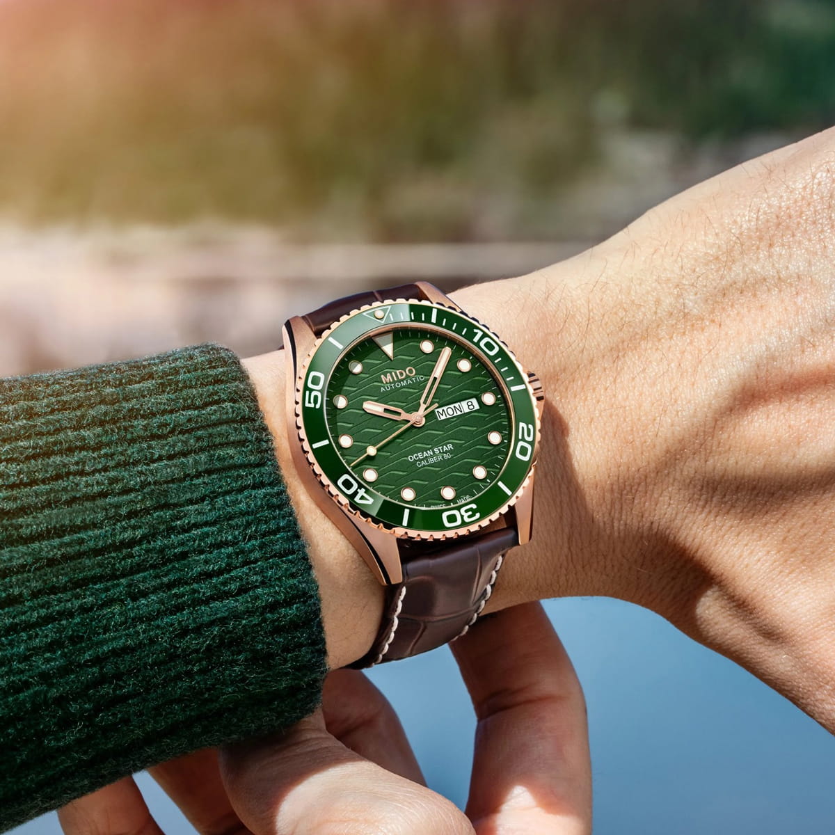Eine Person, die einen grünen Pullover trägt, zeigt eine Armbanduhr mit grünem Zifferblatt, weißen Markierungen, Datumsanzeige und einem braunen Lederarmband. Die Uhr hat ein goldfarbenes Gehäuse und eine Lünette mit weißen Ziffern.
