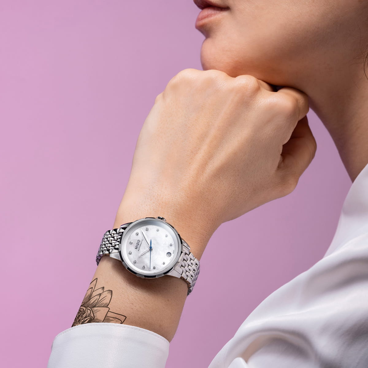 Eine Person, die eine weiße Bluse trägt, stützt ihr Kinn auf ihre Hand und präsentiert eine silberne Mido Rainflower-Armbanduhr und ein Blumen-Tattoo auf ihrem Handgelenk. Der Hintergrund ist hellrosa.