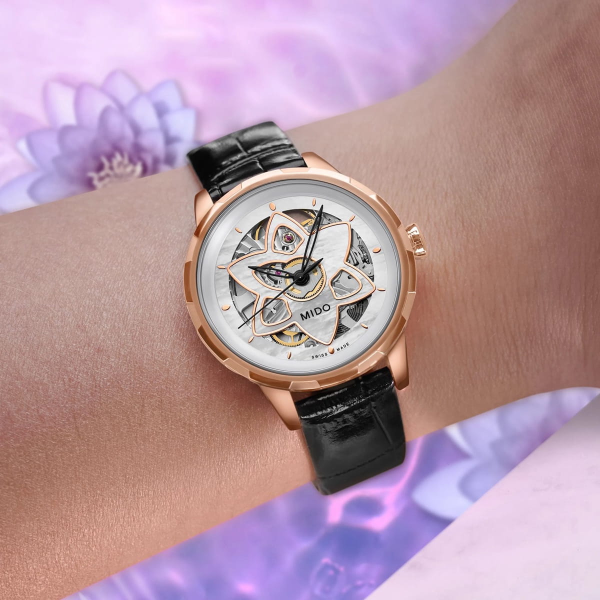 Nahaufnahme eines Handgelenks mit einer roségoldenen Mido Rainflower-Skelettuhr mit weißem Zifferblatt und schwarzem Lederarmband. Der Hintergrund zeigt sanfte Lila-Töne und verschwommene Blumen, die eine elegante, verträumte Atmosphäre schaffen.