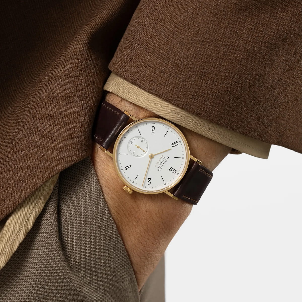 NOMOS Glashütte © 2026 Eine Person, die einen braunen Mantel und eine taupefarbene Hose trägt, hat die Hand in einer Tasche und präsentiert eine klassische NOMOS Glashütte Goldarmbanduhr mit weißem Zifferblatt, schwarzen Ziffern und dunkelbraunem Lederarmband.