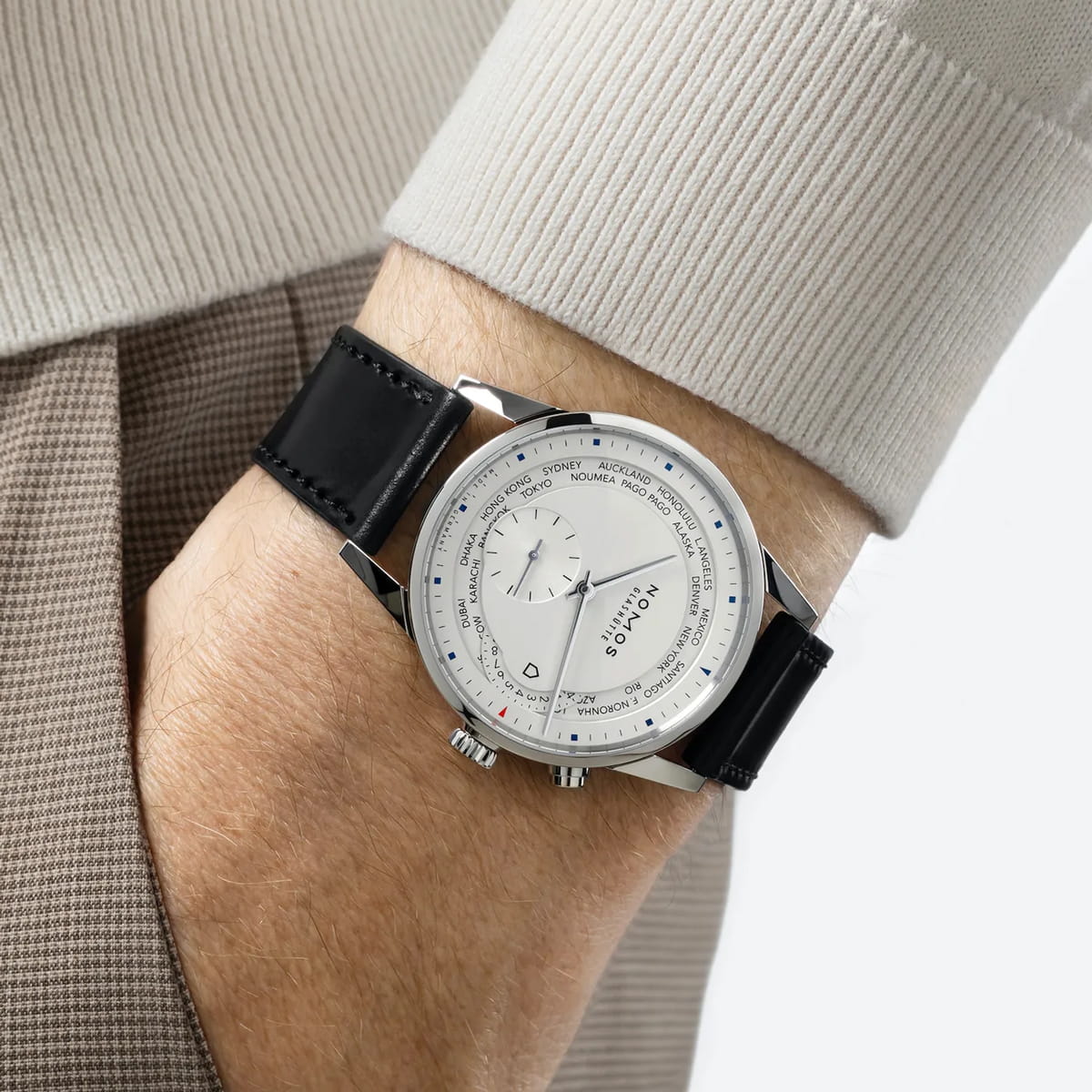 NOMOS Glashütte © 2026 Eine Person, die einen beigen Pullover und eine braune Hose trägt, zeigt eine silberne Armbanduhr von NOMOS Glashütte mit schwarzem Lederarmband und einem weißen Zifferblatt mit mehreren Ziffernblättern und Markierungen.