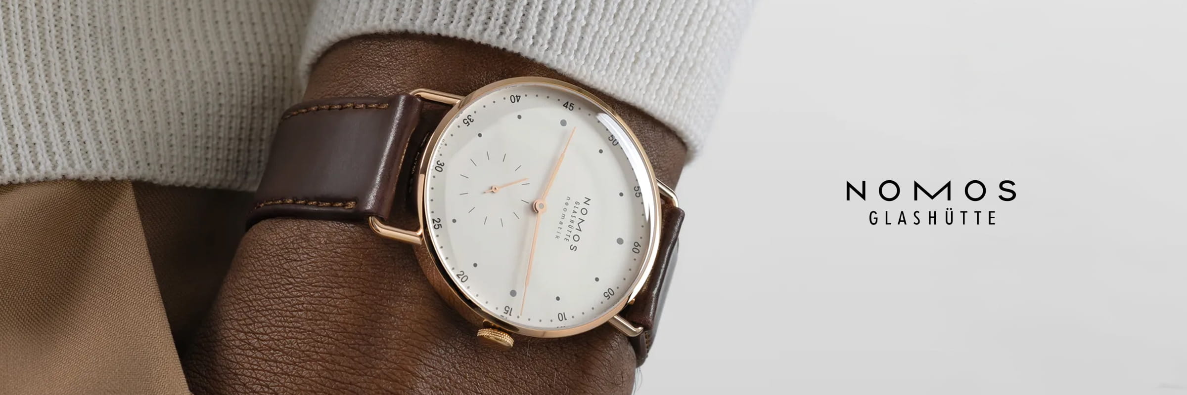 NOMOS Glashütte © 2026 Nahaufnahme einer Person, die eine NOMOS Glashütte-Uhr mit weißem Zifferblatt, roségoldenem Gehäuse und braunem Lederband trägt. Das minimalistische Ziffernblatt zeigt das NOMOS-Glashütte-Logo am rechten Rand des Bildes.