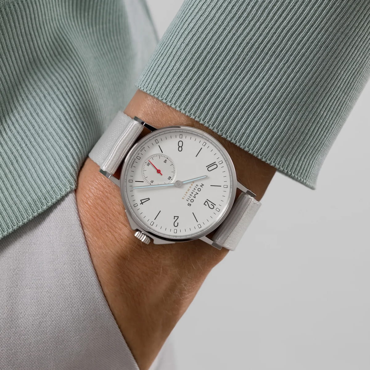 Eine Person trägt eine hellgraue NOMOS-Ahoi-Uhr mit weißem Zifferblatt, schwarzen Ziffern und einem kleinen, rot akzentuierten Hilfszifferblatt. Die Person ist mit einem hellgrünen gerippten Pullover und einer hellgrauen Hose bekleidet.