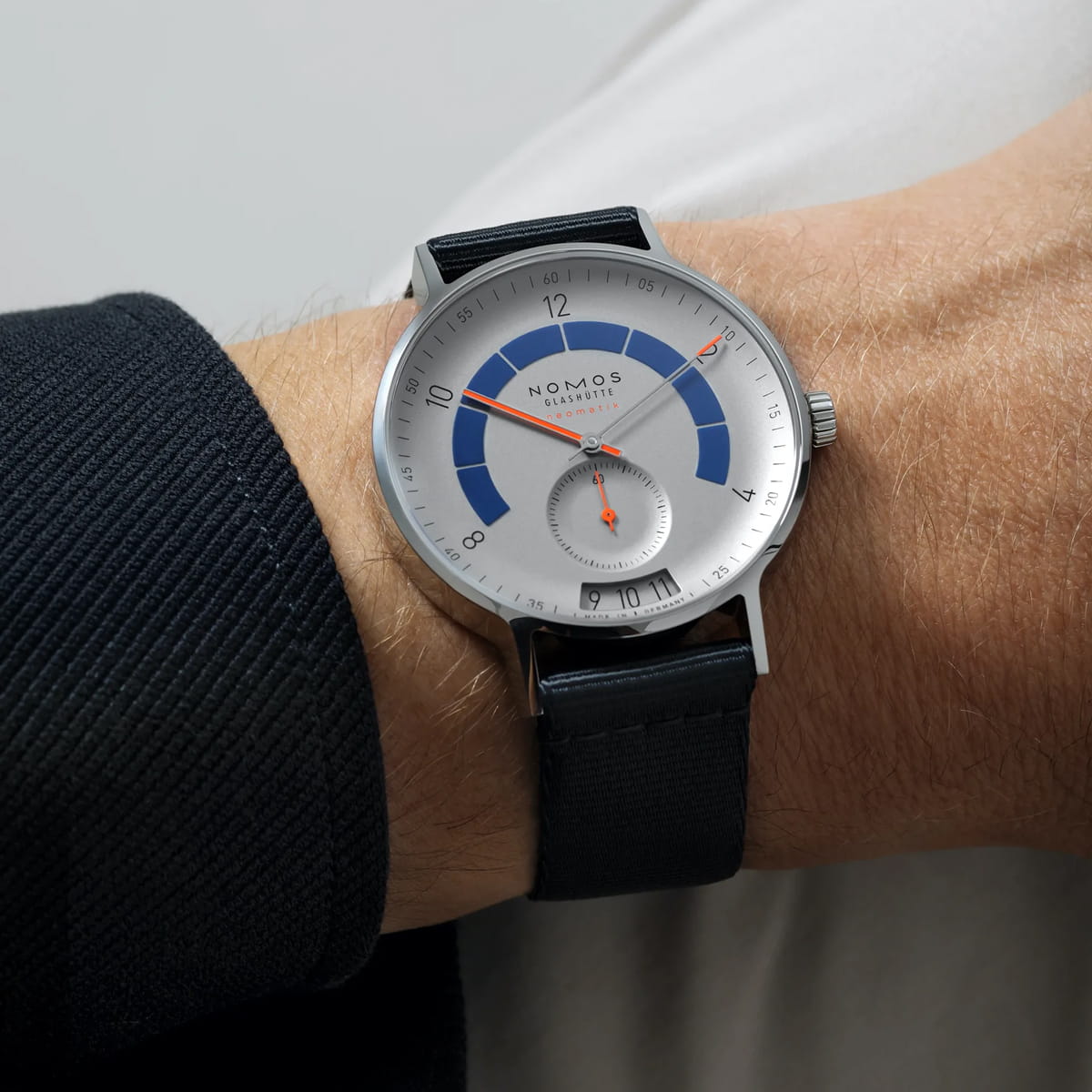 Nahaufnahme eines Handgelenks mit einer NOMOS Autobahn-Armbanduhr mit silbernem Zifferblatt, blauen und orangefarbenen Akzenten, schwarzem Armband und minimalistischem Design. Die Uhr ist mit einer dunklen, strukturierten Hülle kombiniert.