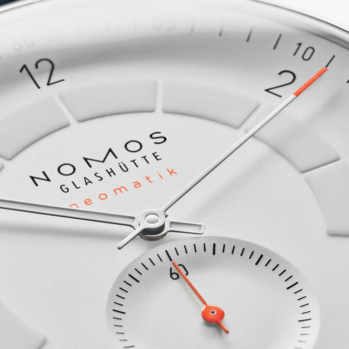 Nahaufnahme eines NOMOS-Glashütte-Zifferblatts, inspiriert von der NOMOS-Autobahn, mit minimalistischem weißem Zifferblatt, schwarzen Ziffern, dünnen silbernen Zeigern und einer kleinen Sekunde auf dem Zifferblatt. Orangefarbene Akzente heben den Sekundenzeiger und den Schriftzug "neomatik" hervor.