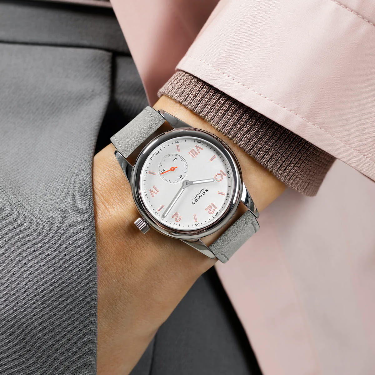Eine Person trägt eine silberne analoge NOMOS-Club-Armbanduhr mit hellgrauem Wildlederarmband und weißem Zifferblatt mit rosafarbenen römischen Ziffern, gekleidet in ein rosa-grau abgestimmtes Outfit.
