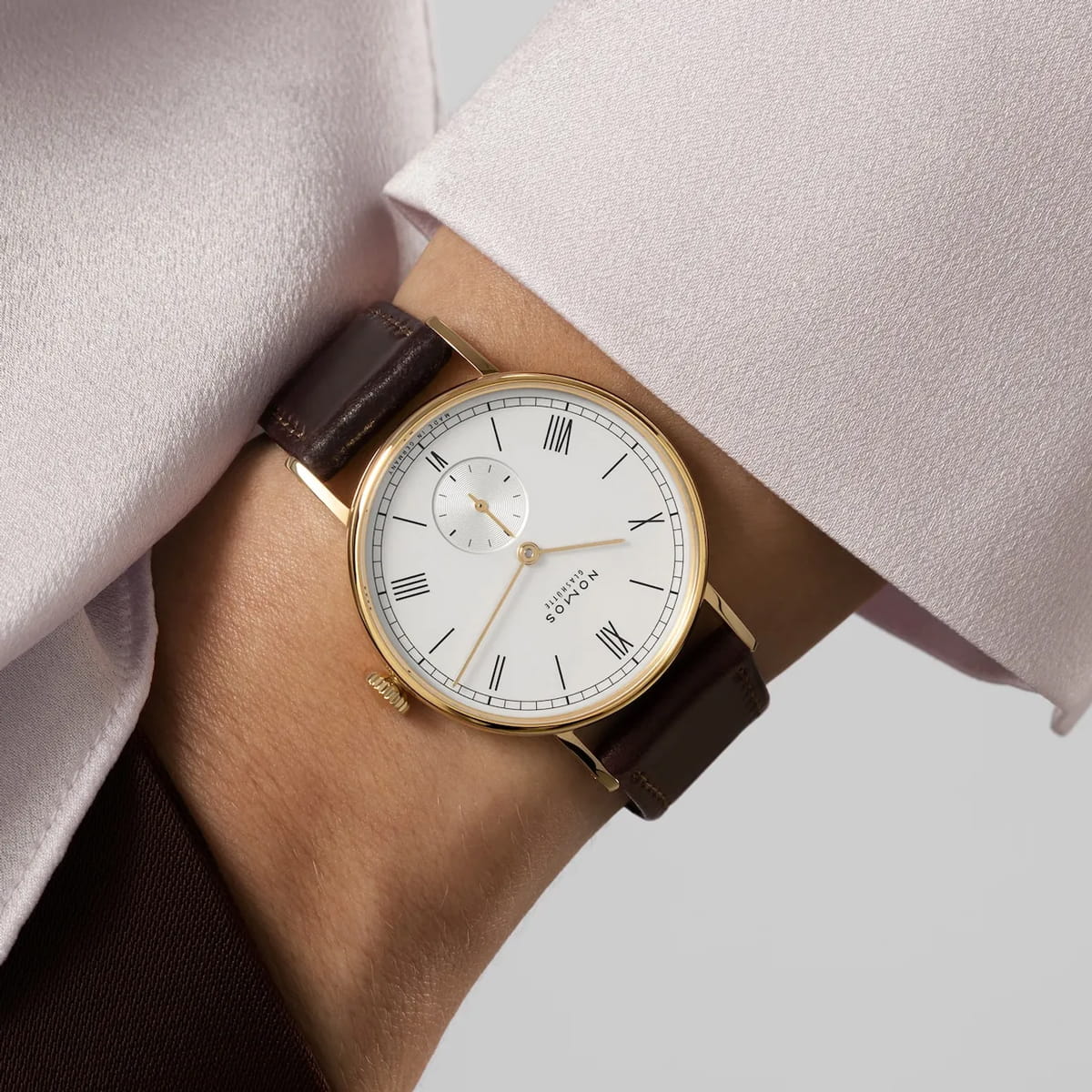 NOMOS Glashütte © 2026 Nahaufnahme des Handgelenks einer Person, die eine goldene NOMOS-Ludwig-Uhr mit weißem Zifferblatt, schwarzen römischen Ziffern, kleiner Sekundenanzeige und braunem Lederarmband trägt, gepaart mit einem blassrosa Langarm.